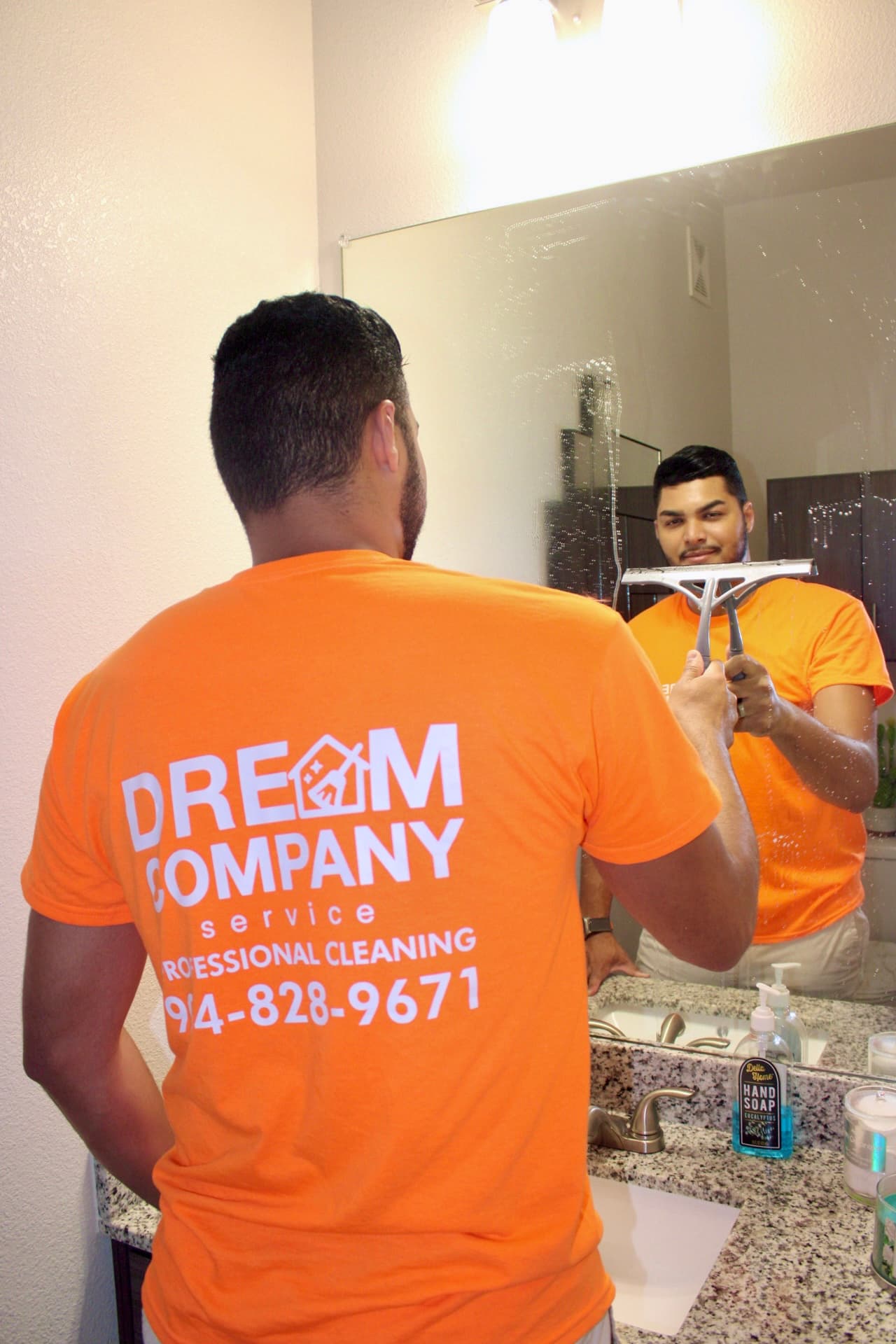Professional cleaner in an orange Dream Company shirt uses a squeegee on a bathroom mirror.
