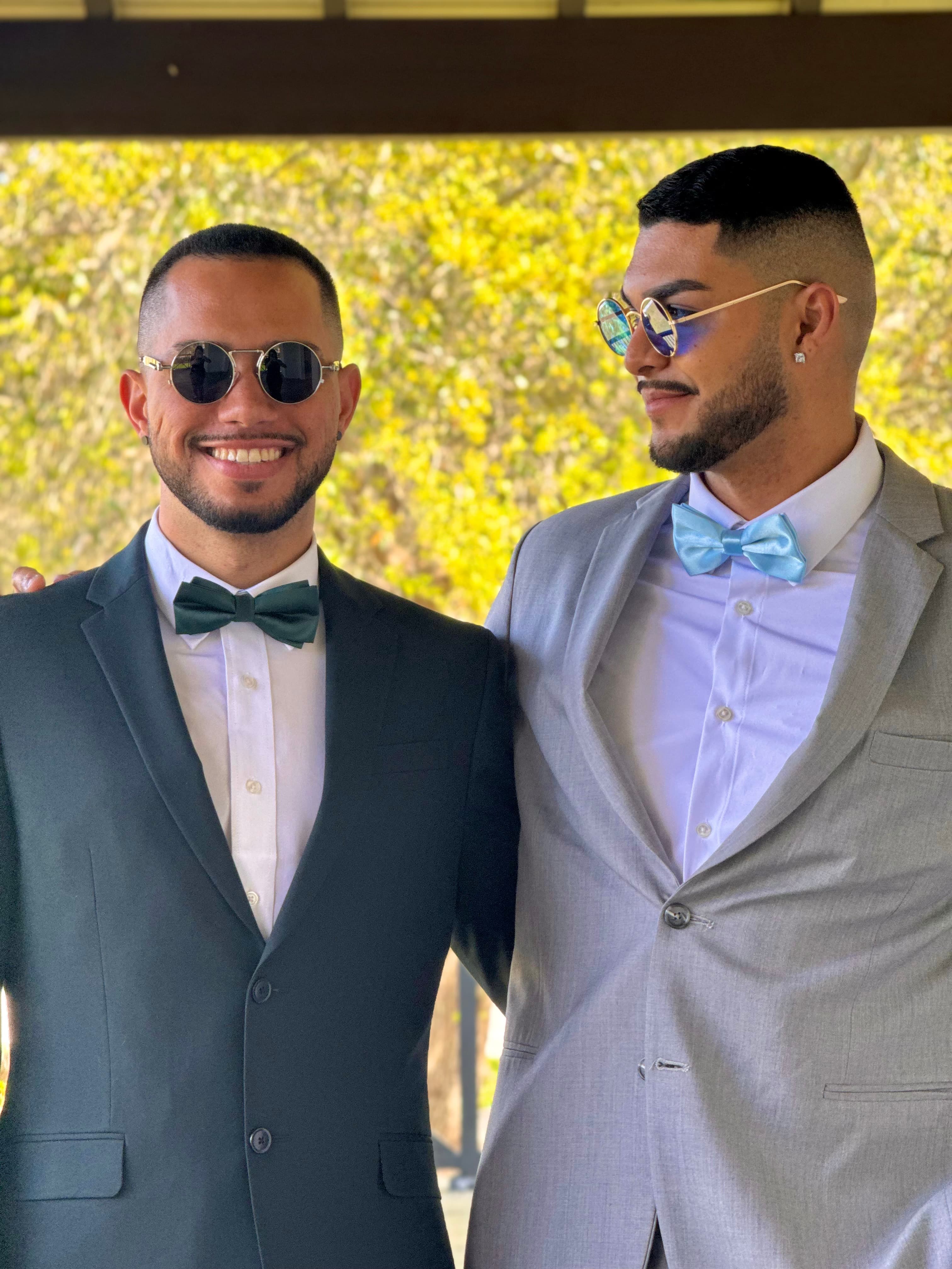 Two men in suits and bowties wearing sunglasses smile together against a leafy background.