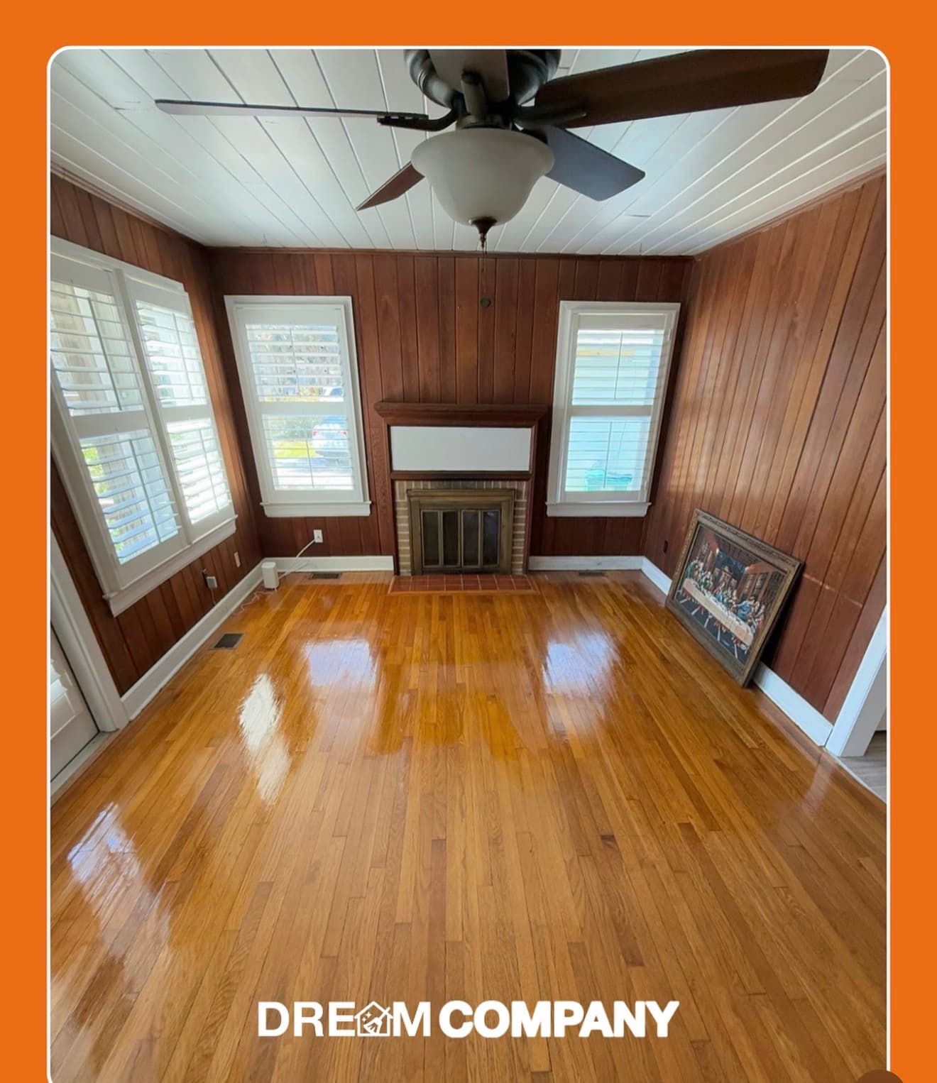 Empty room with polished hardwood floors, wood-paneled walls, a fireplace, and white window shutters.