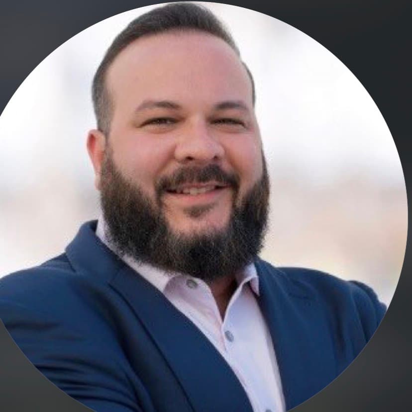 Smiling man with a full dark beard wearing a blue blazer and light pink shirt.